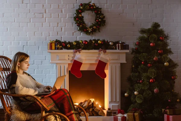 Mujer joven en la mecedora de mimbre usando el ordenador portátil en la sala de estar decorada para la Navidad - foto de stock