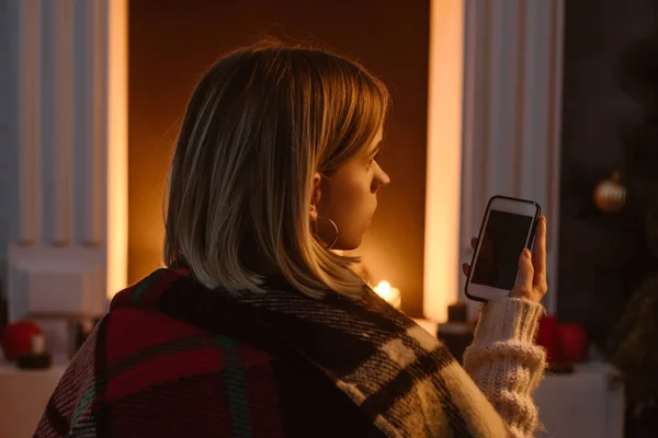 Giovane donna in coperta a scacchi utilizzando smaprtphone con schermo bianco a casa — Foto stock