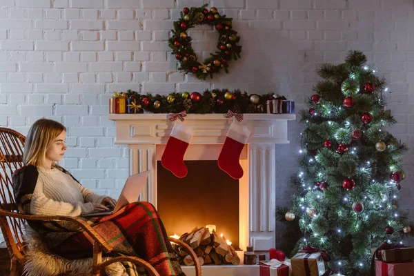 Mujer joven en la mecedora de mimbre usando el ordenador portátil en la sala de estar decorada para la Navidad - foto de stock