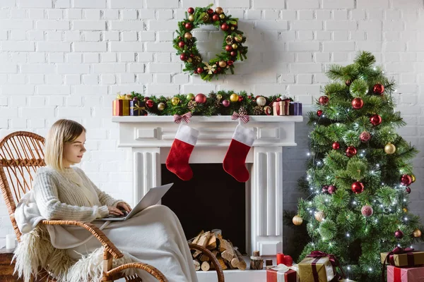 Junge Frau auf Weidenschaukelstuhl mit Laptop im weihnachtlich dekorierten Wohnzimmer — Stockfoto