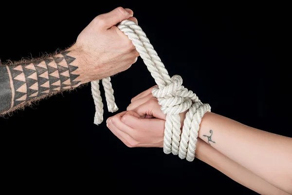 Partial view of man tying rope on female hands with tattoo isolated on black — Stock Photo