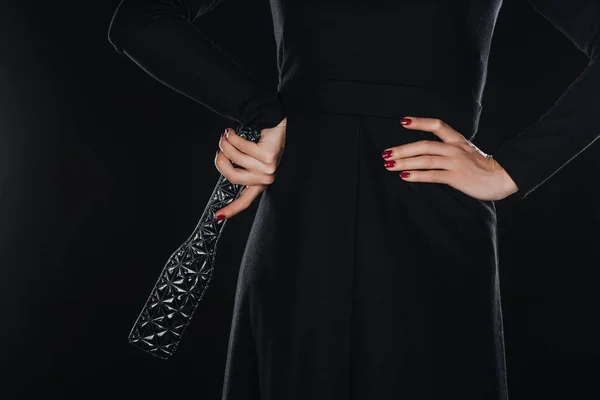 Back view of woman with hand on waist holding leather spanking paddle isolated on black — Stock Photo