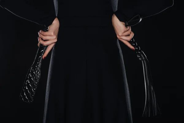 Задний вид женщины с руками на талии держа шлепающую весло и порка кнут изолированы на черном — стоковое фото