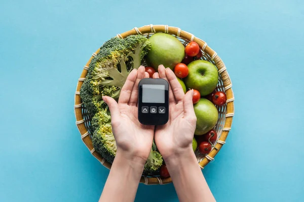Вид зверху на глюкометр у жіночих руках з овочами та яблуками на синьому фоні — стокове фото