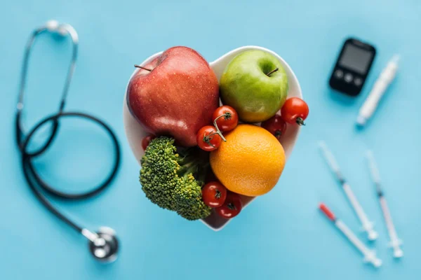 Enfoque selectivo de frutas y verduras en tazón en forma de corazón con equipo médico sobre fondo azul - foto de stock