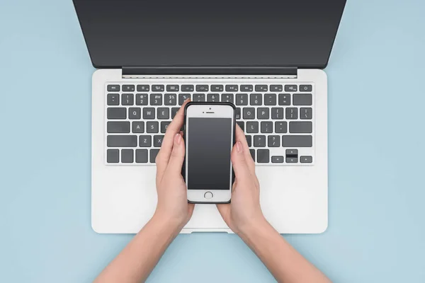 Vista dall'alto della donna che tiene lo smartphone sopra il laptop su sfondo blu chiaro — Foto stock