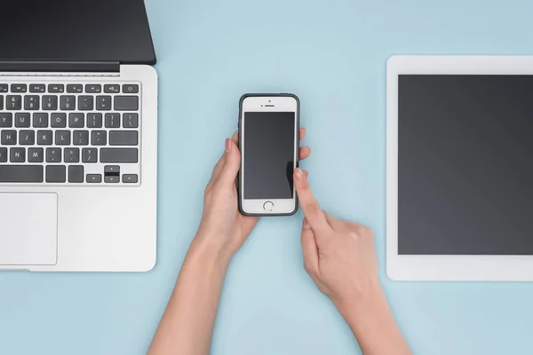 Visão superior da mulher apontando para o smartphone perto de gadgets no fundo azul claro — Fotografia de Stock