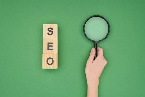 Top view of woman holding magnifying glass near wooden cubes on green background — Stock Photo