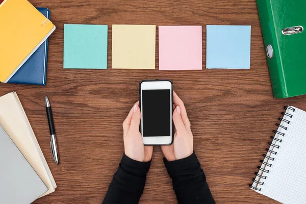 Vista recortada de la mujer sosteniendo teléfono inteligente con pantalla en blanco y cuatro notas adhesivas en el escritorio de oficina de madera - foto de stock