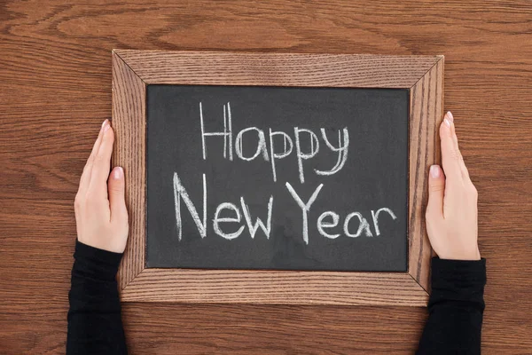 Vista parcial de la mujer sosteniendo pizarra con texto 'feliz año nuevo' sobre fondo de madera - foto de stock