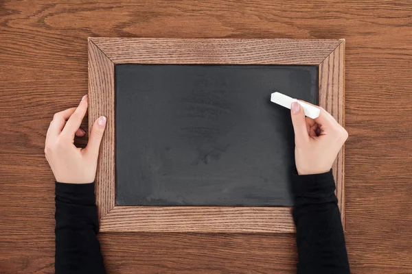 Vista parcial de la mujer sosteniendo un pedazo de tiza sobre una pizarra vacía sobre un fondo de madera - foto de stock