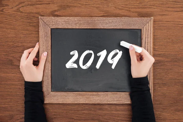 Vista recortada de la mujer sosteniendo tiza y escritura 2019 fecha a bordo con fondo de madera - foto de stock