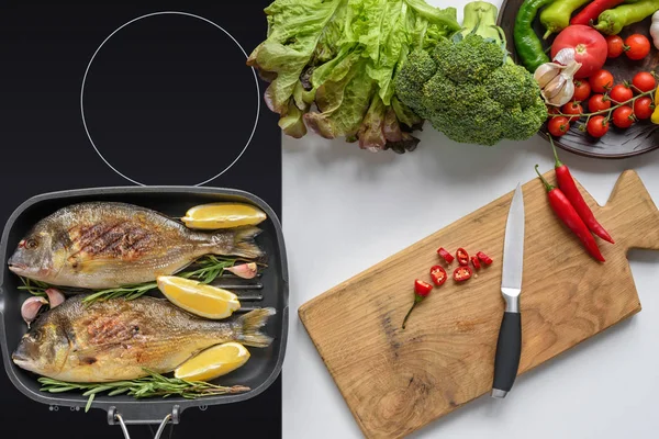 Вид зверху смачна смажена риба на електричній плиті та свіжі овочі з дерев'яною обробною дошкою та ножем — стокове фото