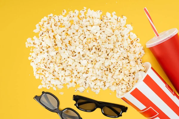 Top view of 3d glasses, red disposable cup with straw and overturned striped bucket with popcorn isolated on yellow — Stock Photo