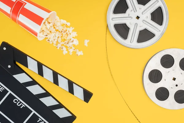 Top view of clapperboard, overturned striped bucket with popcorn and film reels with cinema tape isolated on yellow — Stock Photo