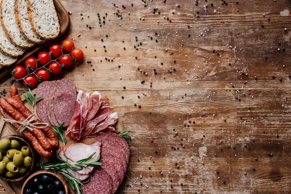 Vista superior de tablas de cortar con sabroso salami, salchichas ahumadas, aceitunas, tomates y pan en la mesa de madera con granos de pimienta dispersos - foto de stock