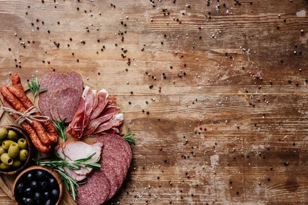 Vue du dessus de la planche à découper ronde avec un savoureux salami, des saucisses fumées et des olives sur une table en bois aux épices éparses — Stock Photo