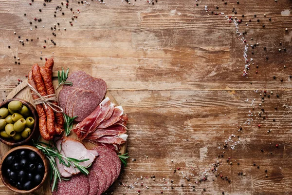 Vista superior da placa de corte redonda com salame saboroso, prosciutto e azeitonas em tigelas em mesa de madeira com especiarias espalhadas — Fotografia de Stock