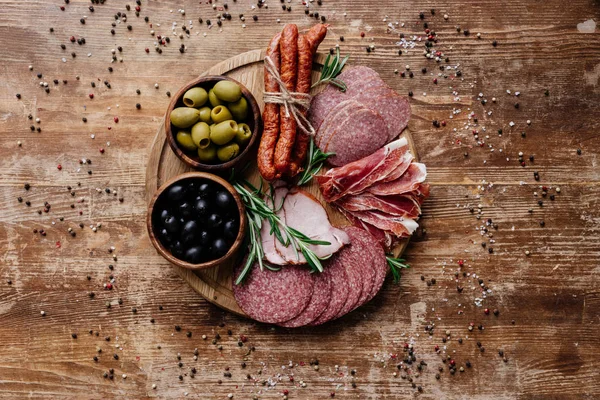 Vista superior de la tabla de cortar redonda con delicioso salami, jamón y aceitunas en dos cuencos en la mesa de madera con especias dispersas - foto de stock