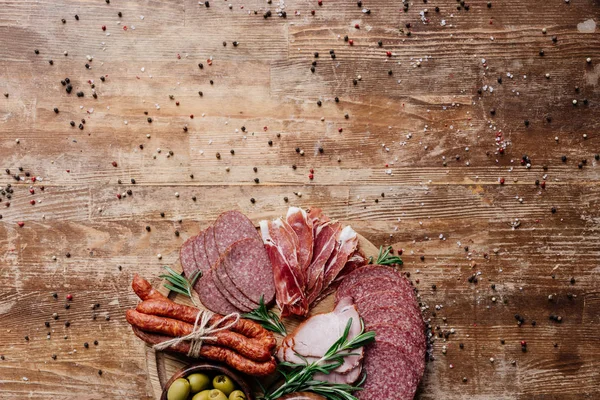 Vista superior da placa de corte com azeitonas e salame fatiado, presunto e presunto em mesa de madeira com especiarias espalhadas — Fotografia de Stock