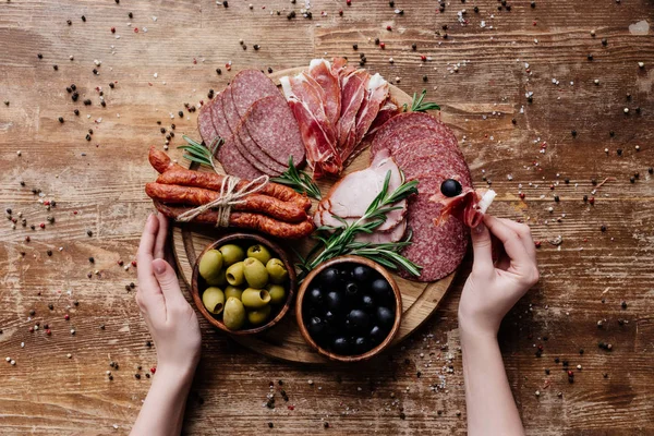 Cropped view of  woman taking prosciutto from round cutting board with olives, salami and ham on wooden table with scattered peppercorns — Stock Photo
