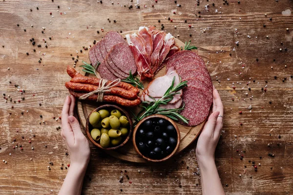 Top view of  female hands holding round cutting board with olives in two bowls and sliced salami, prosciutto and ham on wooden table with scattered peppercorns — Stock Photo