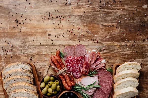 Vista superior de tablas de cortar con delicioso pan, salami, salchichas ahumadas, jamón y aceitunas en mesa de madera con granos de pimienta dispersos - foto de stock