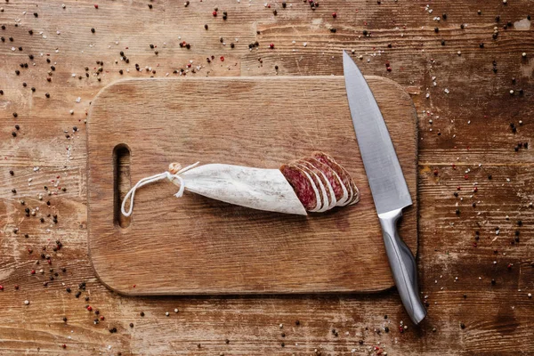 Top view of cutting board with steel knife and tasty sliced salami on wooden table with scattered peppercorns — Stock Photo