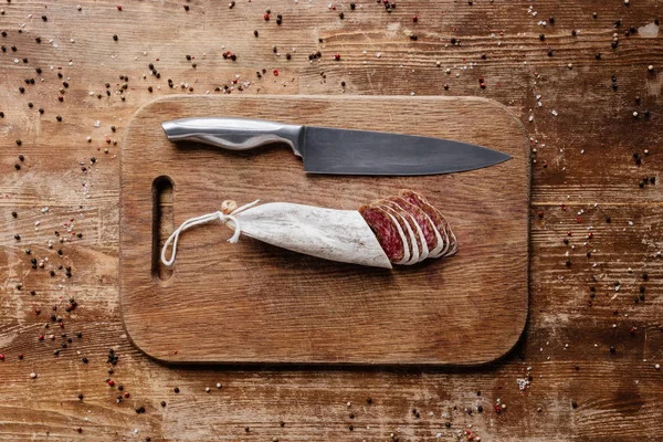 Top view of wooden cutting board with steel knife and sliced salami on table with scattered spices — Stock Photo