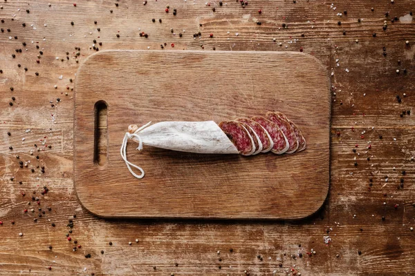 Top view of cutting board with delicious sliced salami on wooden table with scattered peppercorns — Stock Photo