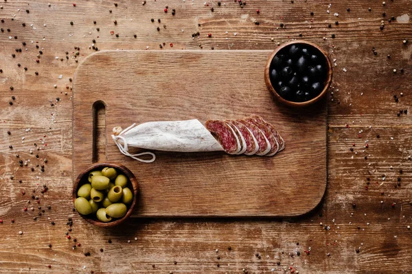 Vista superior de tabla de cortar con aceitunas negras y verdes en dos cuencos y delicioso salami en rodajas en mesa de madera con especias dispersas - foto de stock