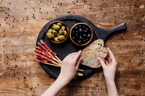 Vista recortada de la mujer poniendo pan en la tabla de cortar redonda con aceitunas en dos tazones y palitos de pan con prosciutto en la mesa de madera - foto de stock