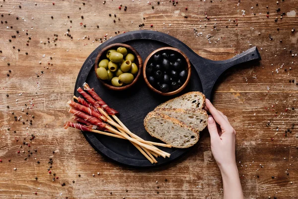 Vista recortada de la mujer poniendo pan en la tabla de cortar redonda con aceitunas, palitos de pan y prosciutto en la mesa de madera - foto de stock