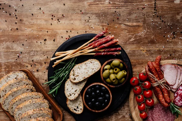Vista superior de tablas de cortar con aceitunas, palitos de pan, jamón, salami, pan, tomates y hierbas en la mesa de madera - foto de stock