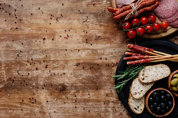 Top view of cutting boards with olives in bowls, breadsticks, prosciutto, smoked sausages, salami, bread, tomatoes and herbs on wooden table — Stock Photo