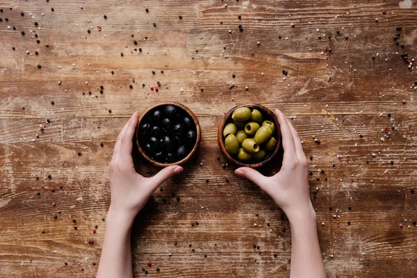 Mains féminines tenant deux bols avec des olives noires et vertes sur une table en bois — Photo de stock