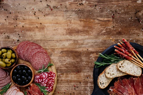 Верхний вид резки досок с оливками, хлебными палочками, прошутто, салями, хлебом и травами на деревянном столе — стоковое фото