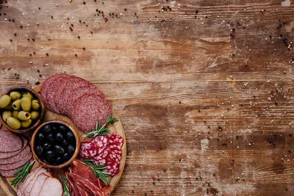 Vue du dessus de la planche à découper avec des olives, des bâtonnets de pain, un délicieux prosciutto, du salami et des herbes sur une table en bois avec des épices éparses — Photo de stock