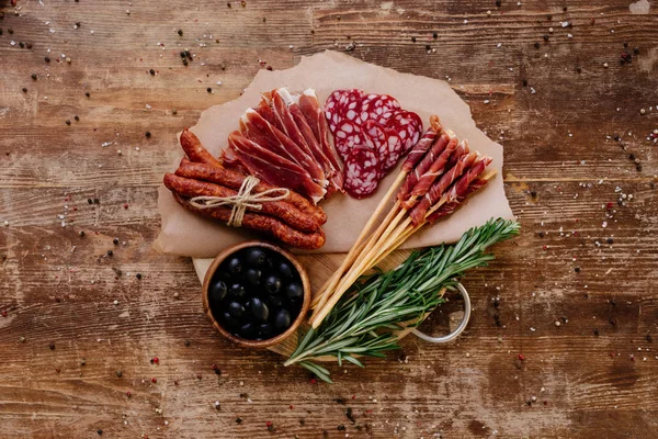 Top view of round cutting board with black olives, delicious prosciutto, salami, smoked sausages and herbs on wooden vintage table with scattered peppercorns — Stock Photo