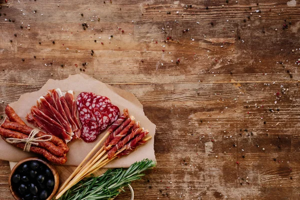 Vue du dessus de la planche à découper ronde avec prosciutto savoureux, salami, saucisses fumées, olives et herbes sur table vintage en bois avec des épices éparses — Photo de stock