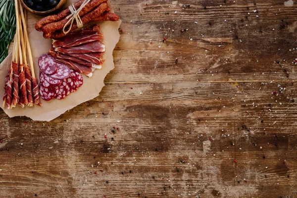Vista superior da placa de corte com prosciutto fatiado, salame e salsichas defumadas em mesa vintage de madeira com especiarias espalhadas — Fotografia de Stock