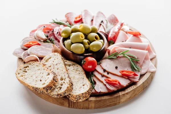 Rundes Schneidebrett mit köstlichen Salami, Schinken, Oliven in Schüssel, Brot, Kräutern und Gemüse auf weißem Tisch — Stockfoto