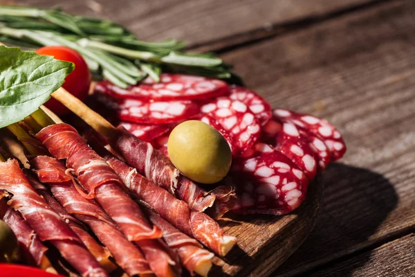 Vista de perto da placa de corte com delicioso salame fatiado, prosciutto e ervas em mesa rústica de madeira — Fotografia de Stock