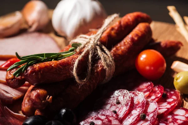 Vista de cerca de la tabla de cortar de madera con delicioso salami en rodajas, salchichas ahumadas, hierbas y verduras - foto de stock