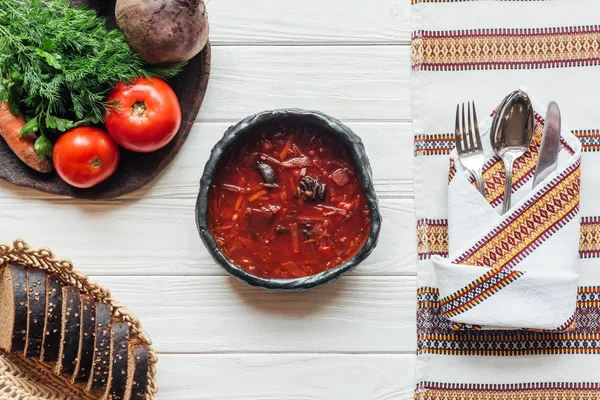 Top view of delicious traditional beetroot soup with cutlery, ingredients and rye bread on white wooden background — Stock Photo