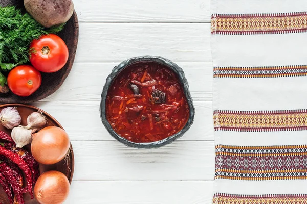 Top view of traditional beetroot soup with ingredients and embroidered towel on white wooden background — Stock Photo