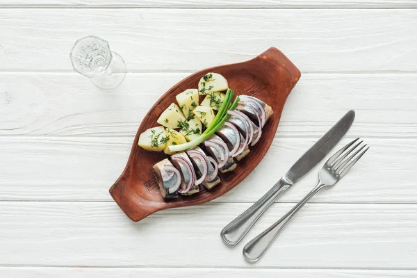 Top view of marinated herring, potatoes and onions in earthenware plate with glass of vodka and cutlery on white wooden background — Stock Photo