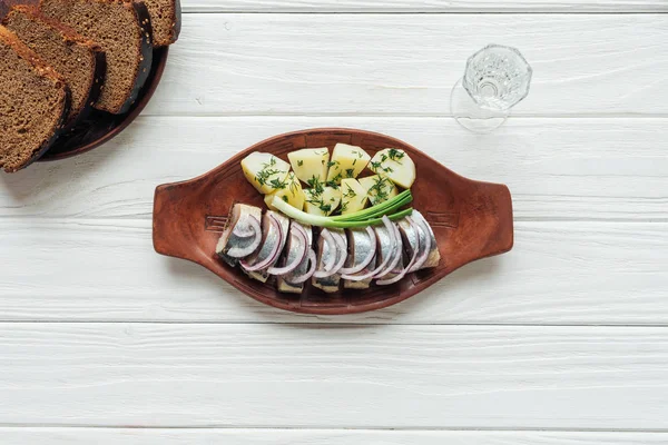 Маринованная сельдь, картофель и лук в глиняной тарелке со стаканом водки и ржаным хлебом на белом деревянном фоне — стоковое фото