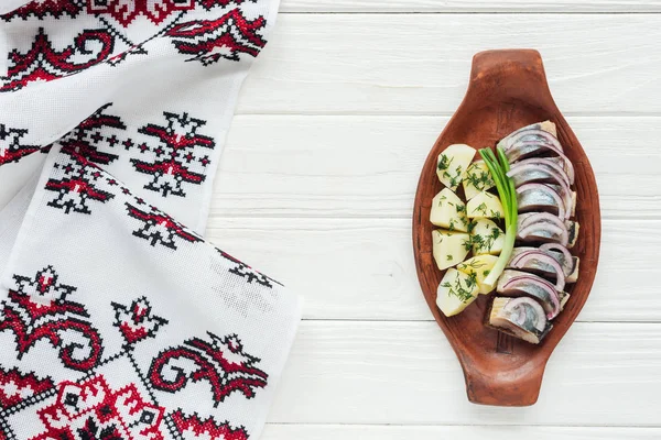 Ansicht von mariniertem Hering mit Kartoffeln und Zwiebeln in Tonteller mit besticktem Handtuch auf weißem Holzhintergrund — Stockfoto