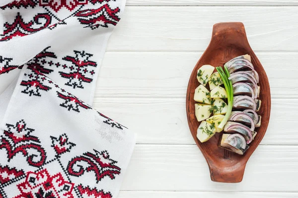 Top view of marinated herring with potatoes and onions in earthenware plate with embroidered towel on white wooden background — Stock Photo